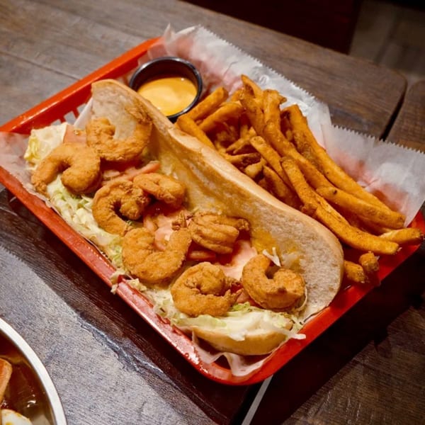 A fried shrimp po'boy sandwich with fries and dipping sauce in a red basket on a wooden table.