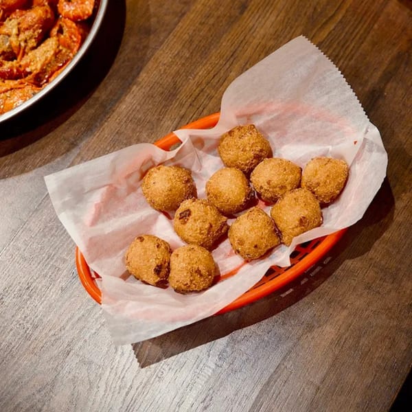 A red basket filled with golden-brown hush puppies on a white paper liner, with a pan of seafood in the background.