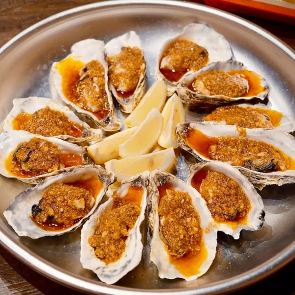 A dozen baked oysters with a savory topping and lemon wedges on a silver platter.